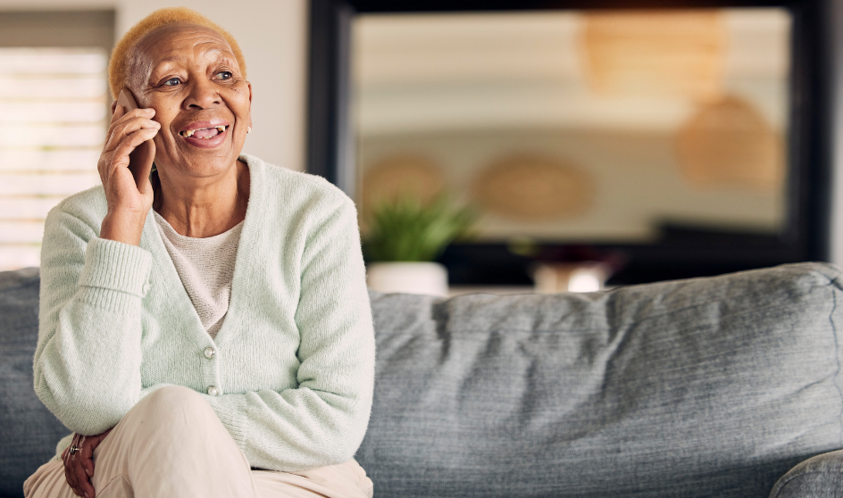 An elderly person smiling while talking on the phone, seated on a grey sofa.