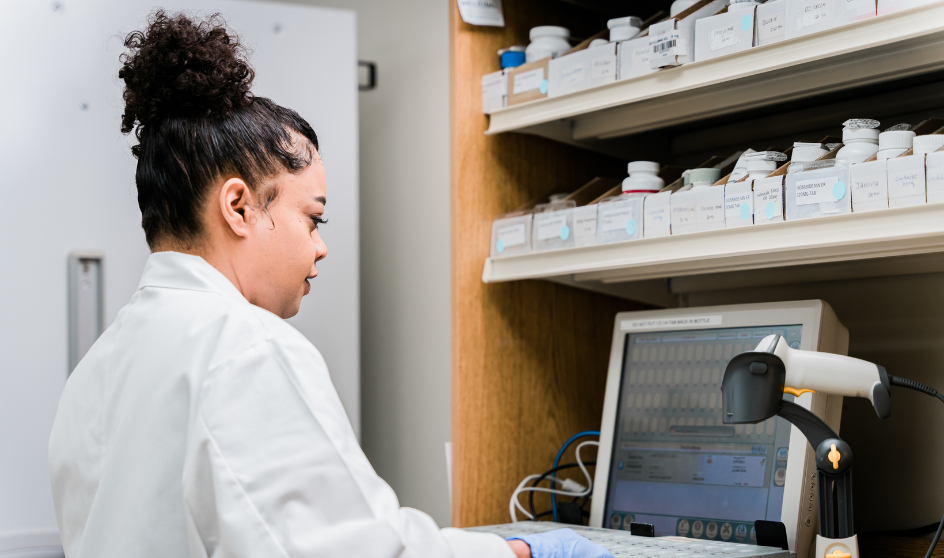 Person in a white lab coat working at a computer with a barcode scanner in a pharmacy or lab setting.