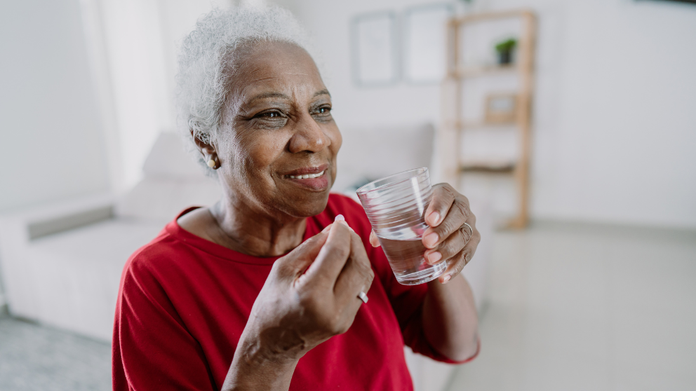 Elderly person in a red shirt holding a pill and a glass of water, smiling in a living room.