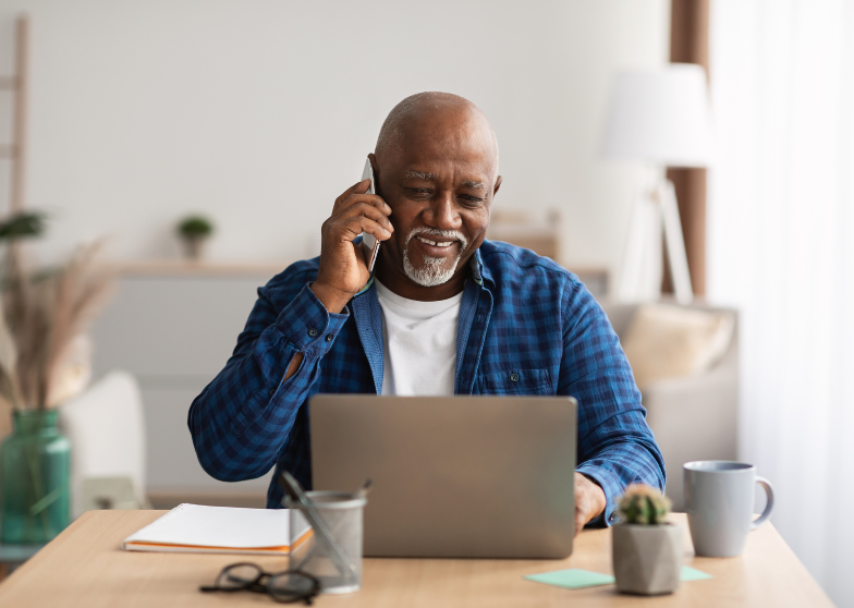 Older man smiling while talking on the phone and using a laptop in a home office.