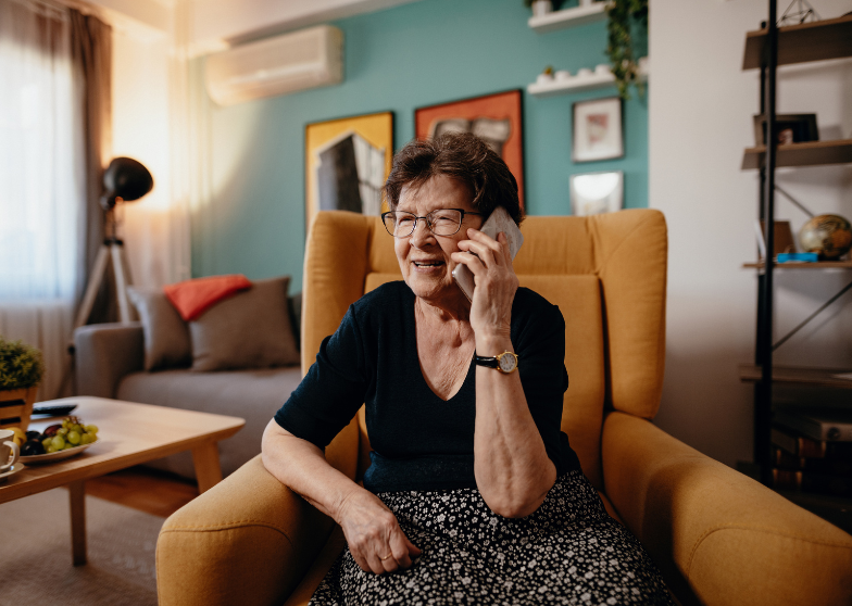 An elderly woman sitting in a yellow armchair, talking on the phone in a cozy living room.